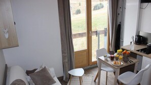 Una habitació acollidora amb un llit, una petita taula de menjador amb cadires, una cuina i una vista a un camp verd a través d'una finestra gran.