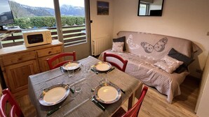 A cozy room with a dining table set for four, a sofa with butterfly-themed bedding, and a view of mountains through large windows. - Ground floor, view ski slopes, balcony, tv, 24m², Le Grand Bornand (Le Grand-Bornand)