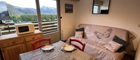 A cozy room with a dining table set for four, a sofa with butterfly-themed bedding, and a view of mountains through large windows.