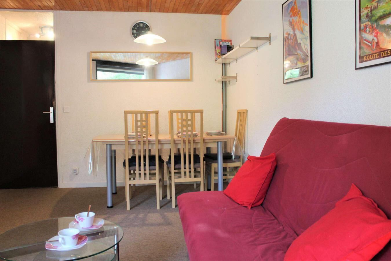 A room with a red sofa, a dining table with chairs, a glass coffee table with cups, a clock, and a mirror on the wall.