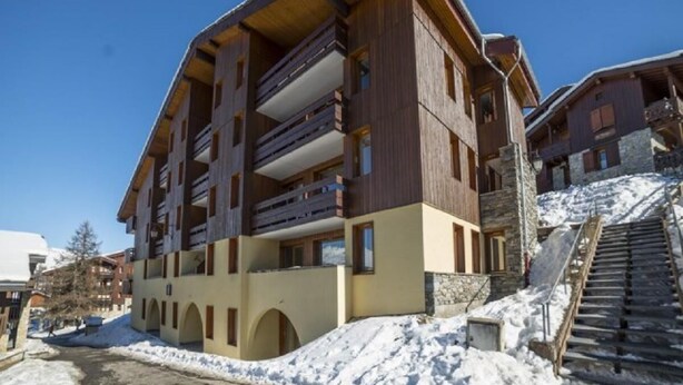 Un hotel de varios pisos con balcones, una pista de esquí al fondo y un cielo despejado.