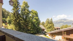 Un albergue de montaña con balcón de piedra, cabañas de madera y vistas del bosque y montañas circundantes.