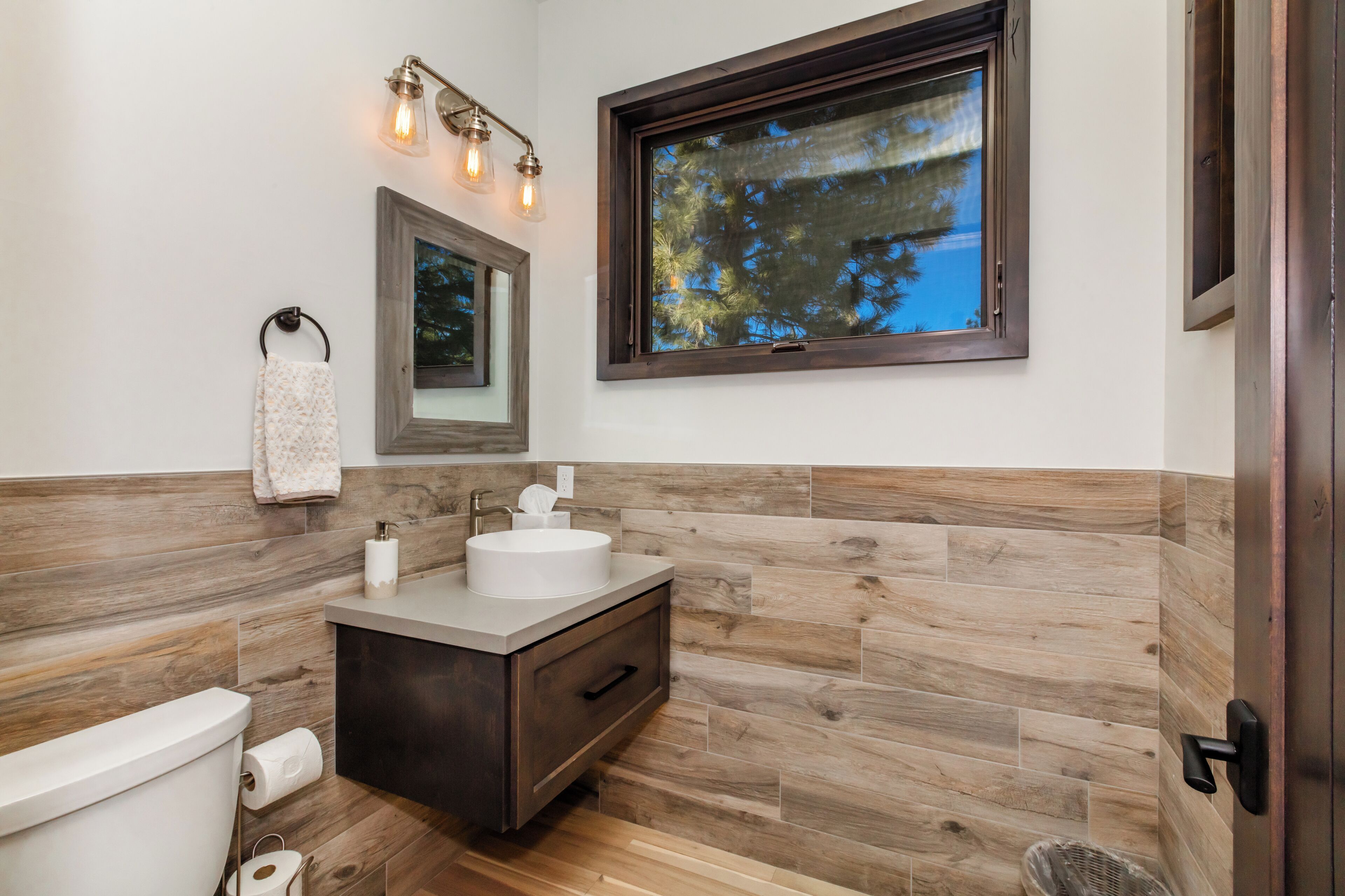 A modern bathroom with a wooden wall backsplash, a large window, and a vanity with a sink and mirror.