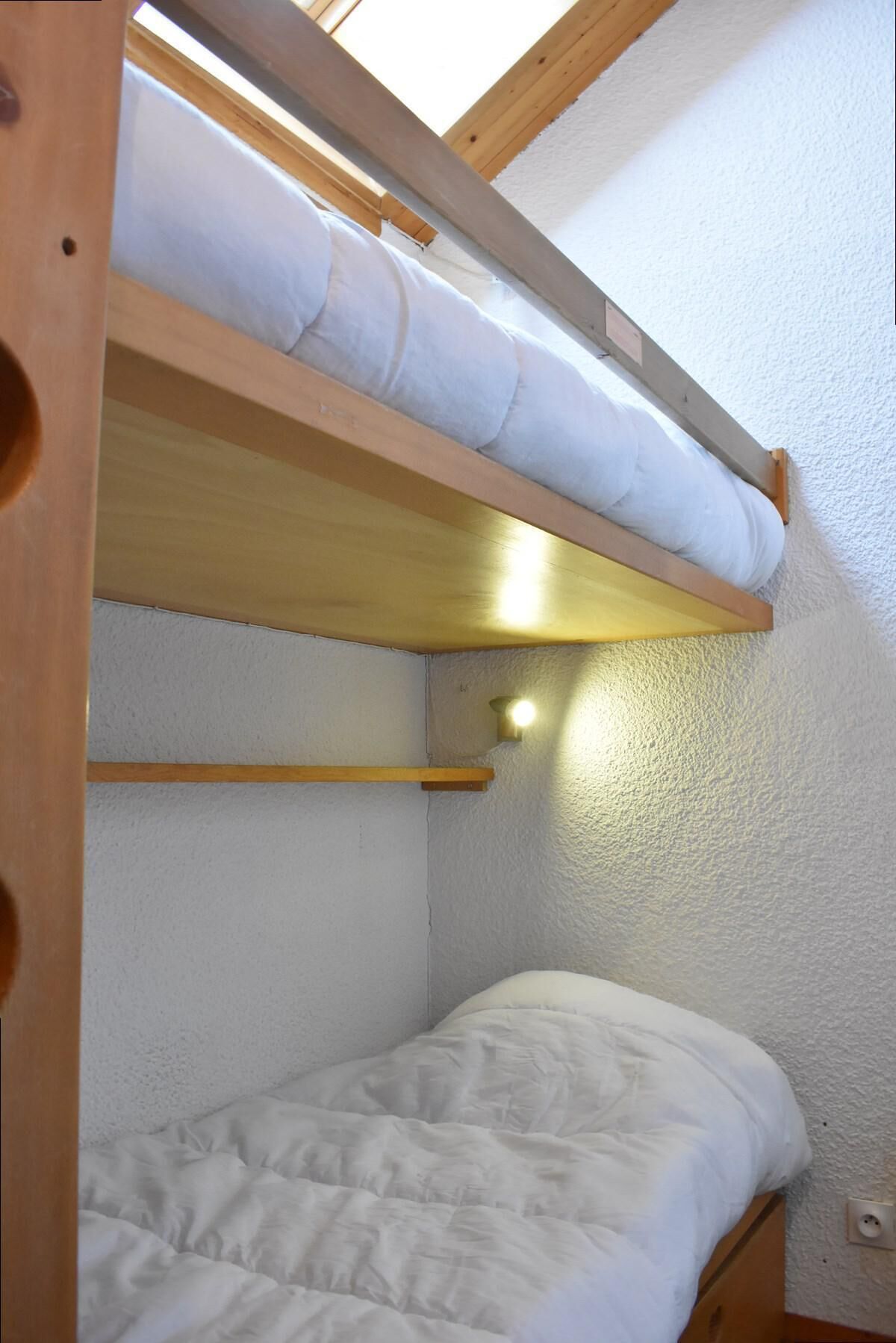 A bunk bed with a skylight above, a single bed below, and a small shelf.