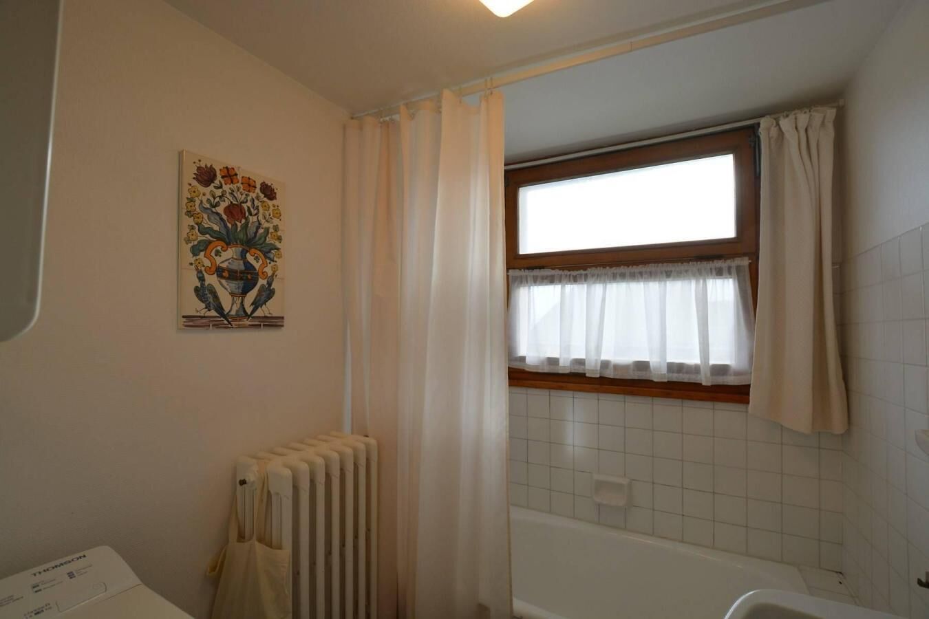 Ein Badezimmer mit Heizkörper, Fenster mit Vorhängen und einer gefliesten Wand.