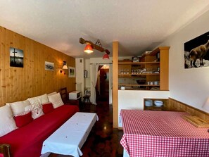 Una habitación acogedora con un sofá rojo, una mesita con mantel a cuadros, estanterías de madera con vajilla y una lámpara de pared.