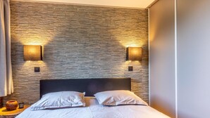 Un dormitorio moderno con pared de diseño que imita piedra, dos lámparas de noche, una cama con ropa de cama blanca y una mesita con un florero y una vela.