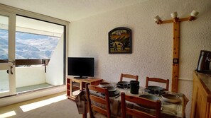 Uma área de refeições com uma mesa posta para quatro pessoas, uma televisão sobre um suporte e vista para montanhas nevadas através de uma janela grande.