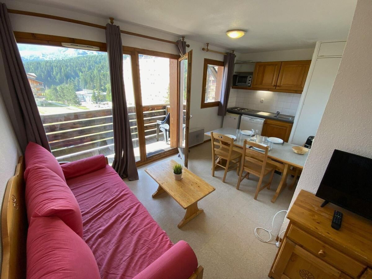 A room with a sofa, a dining table with chairs, a kitchen area, and a balcony with curtains.