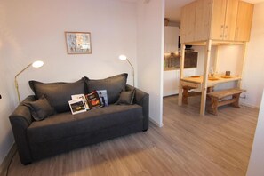 Una sala moderna con un sofá gris oscuro, dos lámparas de pie y revistas. En el fondo, una mesa de comedor de madera y un banco.