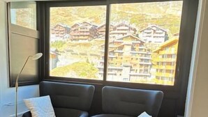 Een moderne kamer met twee zwarte fauteuils en een bijpassend pouf, een vloerlamp en een groot raam met uitzicht op een woonwijk.
