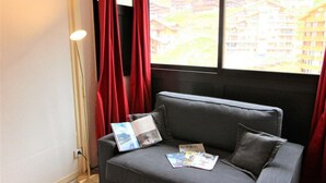 Een grijze bank met een tijdschrift en een kussen, een vloerlamp en een raam met rode gordijnen.