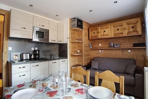 Uma cozinha compacta com micro-ondas, máquina de café e armários de madeira. Uma mesa de jantar com cadeiras para dois, e um sofá ao fundo.