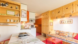 Een gezellige kamer met een eettafel gedekt voor twee personen, een bank met een kleurrijk kussen, houten kasten met een nummer, en een magnetron op het aanrecht.