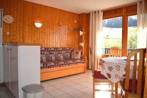 A cozy room with a wooden wall, a sofa, a dining table with chairs, and a window with a view of greenery. - 50m from resort center, floor -1, view mountain, balcony, tv, 31m², Le Grand Bornand (Le Grand-Bornand)