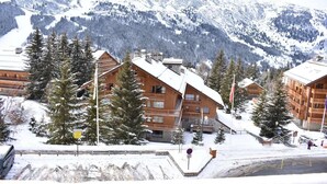Un chalet de montagne enneigé, composé de plusieurs bâtiments en bois, entouré d’arbres et de montagnes couverts de neige.