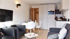 Un salon moderne avec un téléviseur, un coin repas avec une table et des chaises, et une cuisine équipée d’une machine à café.