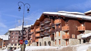 A multi-story hotel with stone and wooden balconies, a ski slope in the background, and a clear blue sky. - 100m from ski slopes, 1st floor, balcony, tv, 23m², La Plagne (La Plagne)