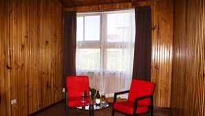 Una habitación con paredes de madera y un amplio ventanal con cortinas. Hay dos sillas rojas y una mesita con una botella y vasos.