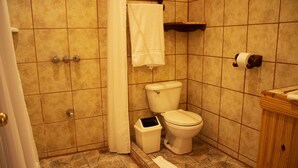 Un baño con inodoro, un cubo de basura, cortina de ducha y estante de madera.