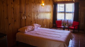 Una habitación con paneles de madera, una cama, dos sillas rojas, una mesita y una ventana con cortinas.