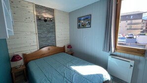 Ein Schlafzimmer mit einem hölzernen Bett, zwei Nachttischen mit Lampen, einem Heizkörper und einem Fenster mit Blick auf parkende Autos und Gebäude.