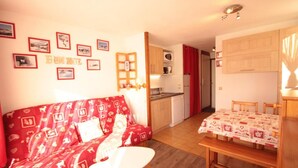 Un salon chaleureux avec un canapé rouge, une table à manger recouverte d’une courtepointe et un coin cuisine doté de meubles en bois et d’un rideau rouge.