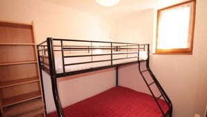 Un lit superposé avec un matelas rouge, une étagère en bois et une fenêtre laissant entrer la lumière.