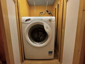 A compact, white front-loading washing machine in a narrow, wooden-walled laundry room with a yellow wall and a ceiling vent.