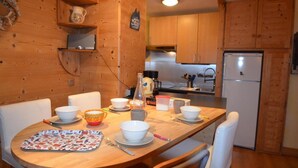Una mesa de comedor de madera con sillas blancas, rodeada de cuencos y platos. Se ve en el fondo una zona de cocina con armarios de madera y un refrigerador.