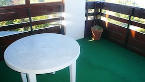 Una mesa redonda blanca con cuatro patas sobre un balcón alfombrado de verde, con barandillas de madera y una planta en maceta.