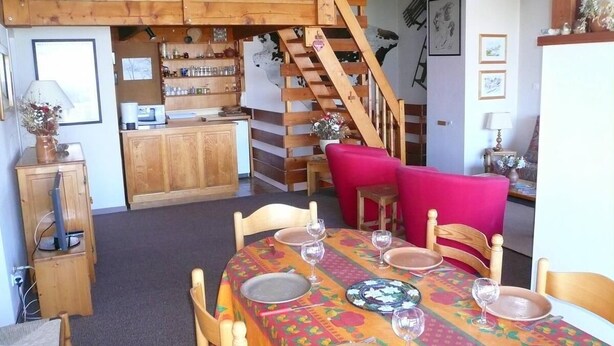 Comedor con escalera de madera, cocina al fondo y una mesa de comedor dispuesta con platos y vasos.