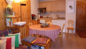 A cozy living room with a round table, wooden chairs, a sofa with patterned cushions, and a kitchen area with wooden cabinets and a microwave.