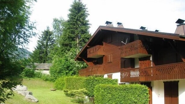 A two-story wooden chalet with a balcony, surrounded by a well-maintained garden and trees.