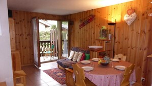A cozy wooden interior with a dining table set for four, a sofa, and a sliding glass door leading to a balcony.