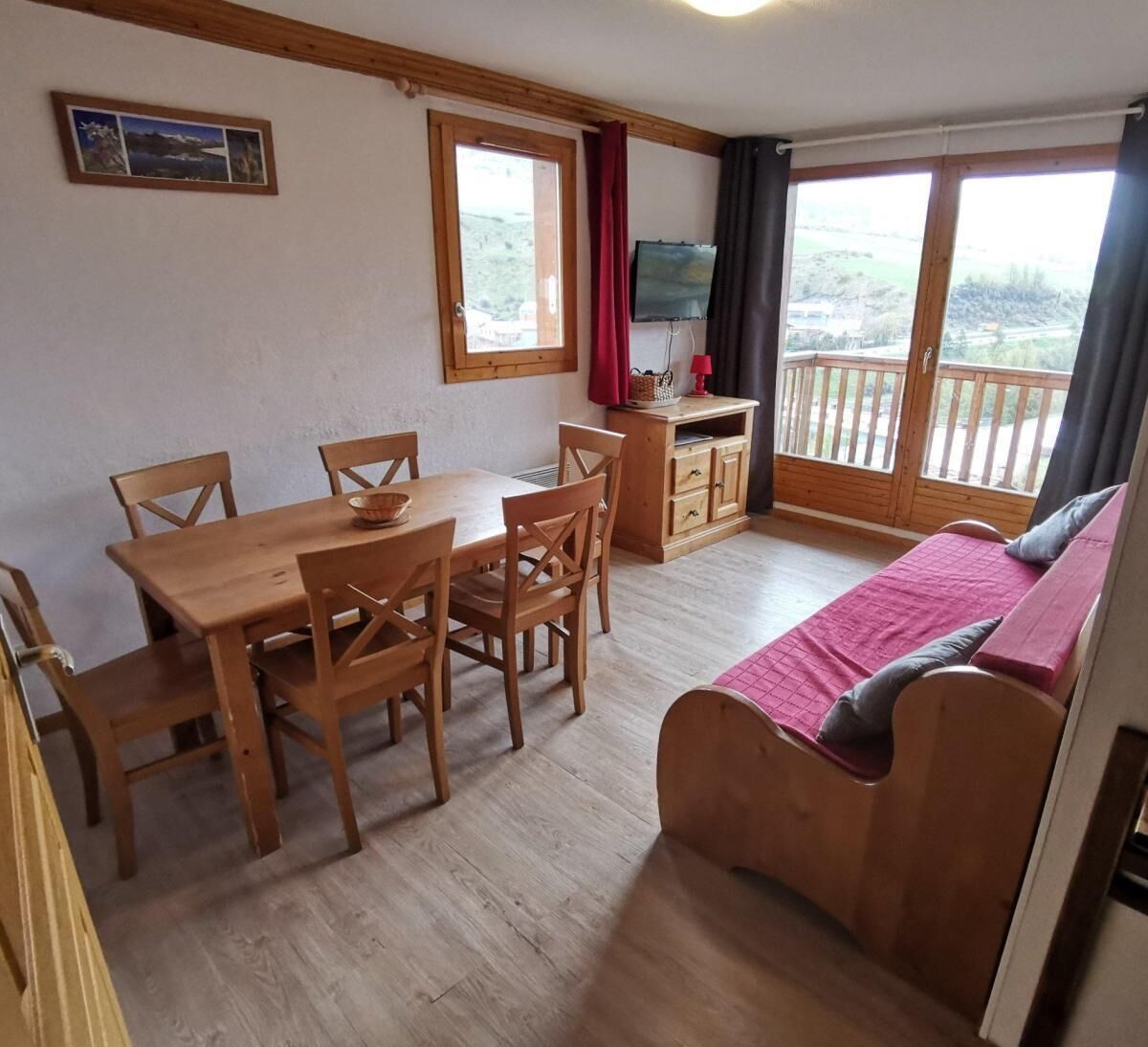 A room with a wooden dining table, chairs, a sofa, a TV, and a balcony with a view.