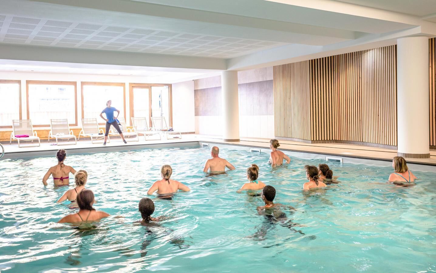 Une piscine avec des personnes dans l’eau, une personne qui passe, et une vue sur l’espace piscine avec des chaises et un mur en bois.