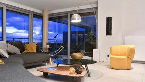 Un soggiorno moderno con un divano, una poltrona gialla, un tavolino in vetro e vista sulla città al crepuscolo.