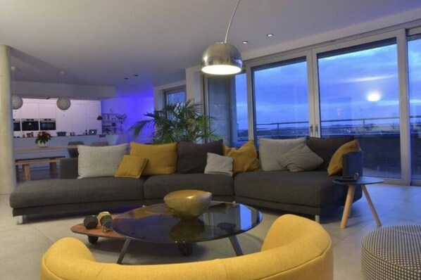 A modern living room with a large sofa, a glass coffee table, and a view of the city through large windows.