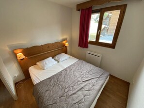 A single bed with a headboard, bedside lamps, a window with a red curtain, and a radiator.