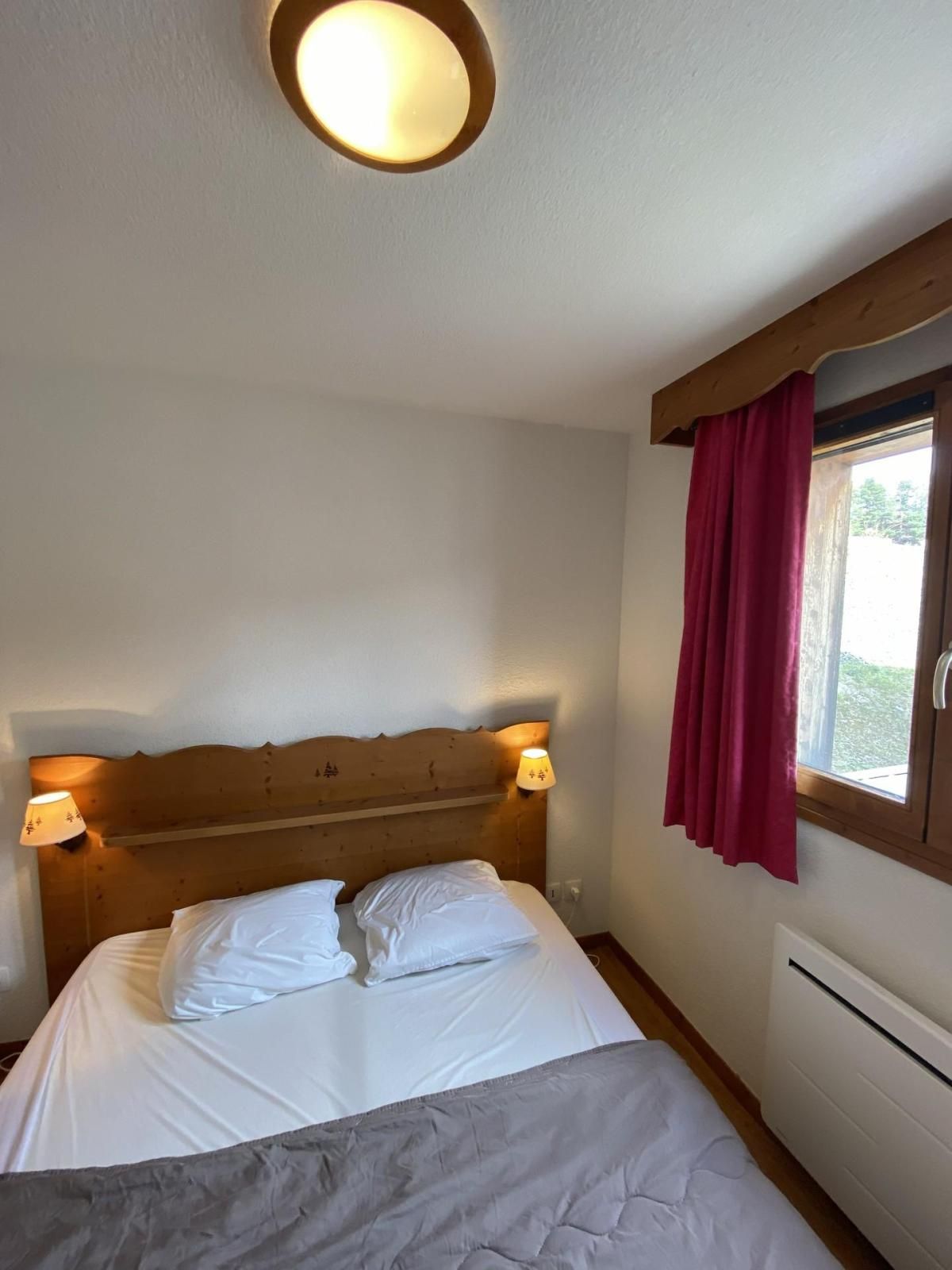 Ett sovrum med en sänggavel i trä, två sänglampor, en säng med vita sängkläder, ett fönster med en röd gardin och en inbyggd luftkonditioneringsanläggning.