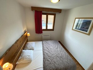 A bedroom with a wooden headboard, a bed with white linens, a window with a red curtain, a radiator, and a framed picture on the wall.