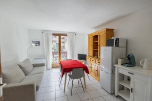 A compact living space with a kitchenette, a dining area with a red table, a sofa, and a balcony with curtains.