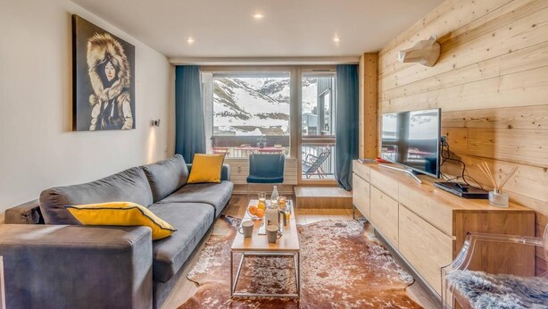Ein modernes Wohnzimmer mit einer Couch, einem Couchtisch und einem Fernseher. Ein großes Fenster bietet Blick auf eine verschneite Landschaft.