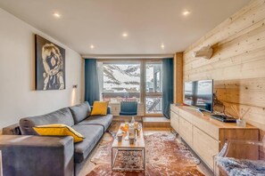 A modern living room with a sofa, coffee table, and a TV. There is a large window with a view of a snowy landscape.