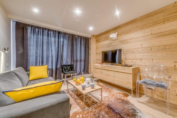 A modern living room with a grey sofa, yellow cushions, a coffee table with fruit and a mug, a TV on a wooden wall, and a clear chair.