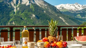 Mesa dispuesta con frutas, una botella de vino y copas, con vistas a un paisaje montañoso.