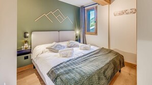Una cama bien hecha con sábanas blancas y una manta de diseño gris y blanco, una mesita de noche con una botella y vasos, una lámpara de pared y una ventana con vistas a la vegetación.