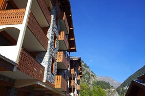 A multi-story building with balconies, stone facade, and a mountainous backdrop.