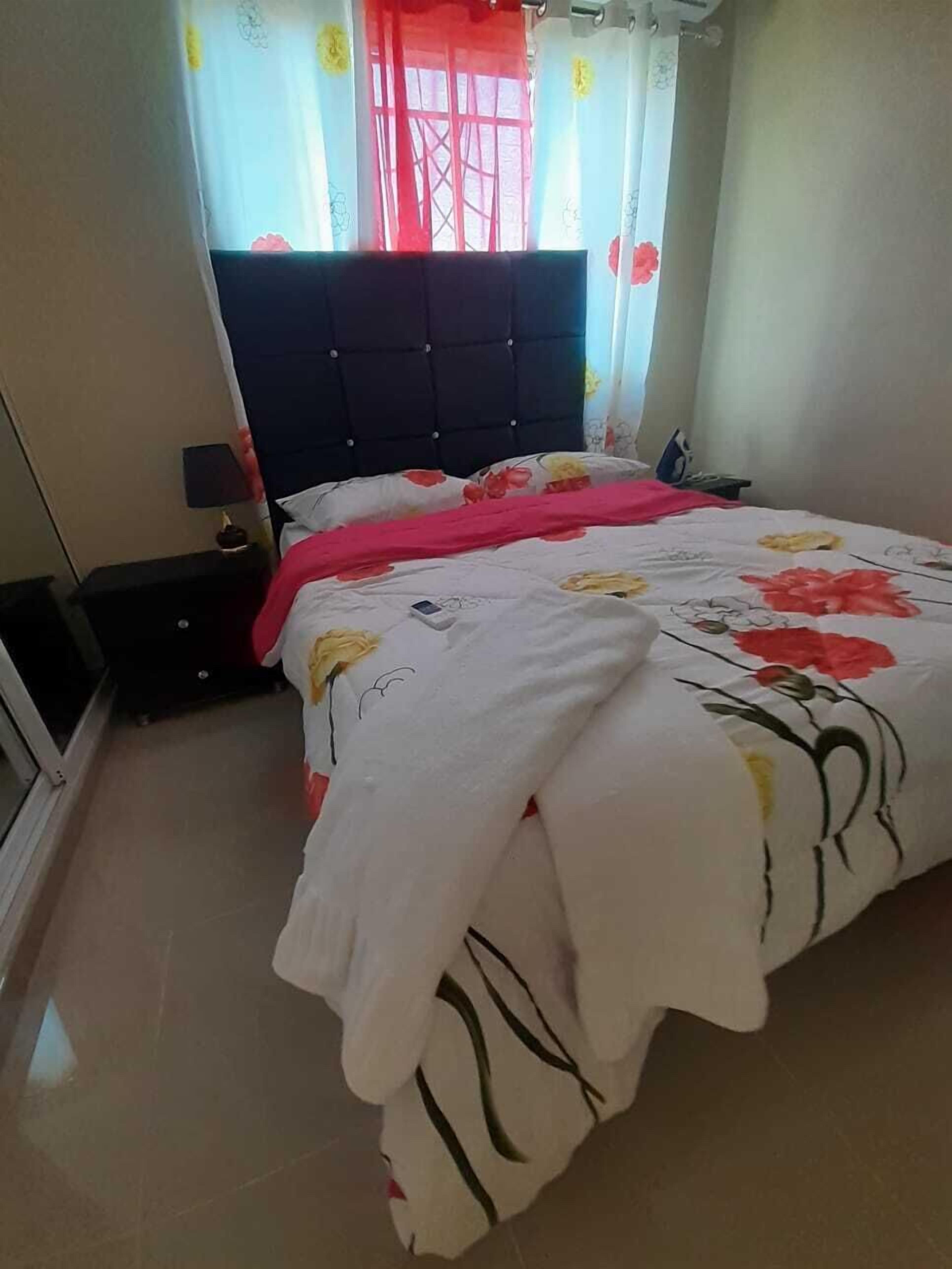 A bedroom with a bed, a nightstand, a window with curtains, and a wall-mounted heater.